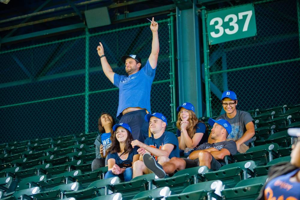 A group of fans wearing the blue gvsu night tigers hat sits near the top row of the stadium. One of the group is standing, fingers pointed at the sky, cheering for the tigers